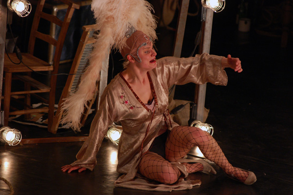 Performer in elaborate costume sits on stage, illuminated by dramatic lighting. Wear cross-legged, with a feathered headdress and satin robe.