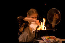 A woman in patterned clothing and apron looks tense as she holds a paper above a flaming pot on a stove. Bright flames and sparks fly.
