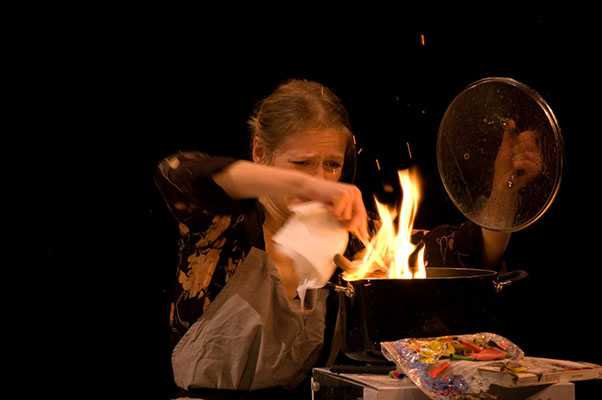 A woman in patterned clothing and apron looks tense as she holds a paper above a flaming pot on a stove. Bright flames and sparks fly.