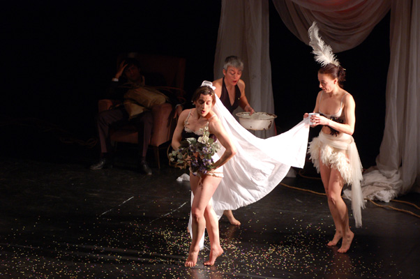 A bridal-themed performance with three women on stage. One wears a veil and holds flowers, another assists with the veil, and a seated man appears contemplative. The mood is theatrical.