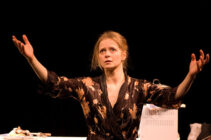A woman stands on stage wearing a dark floral dress, arms raised expressively. Her face shows a mix of determination and concern, set against a dark background.