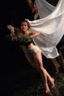 A dancer in beige shorts energetically poses on stage holding a white sheet and leafy branches, conveying a sense of movement and grace.