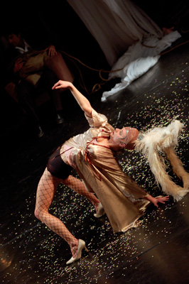 A performer in fishnet tights and a flowing metallic outfit leans back dramatically, with confetti scattered on the dark stage, creating a dynamic and theatrical scene.
