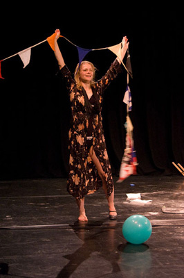 A person on stage, smiling with arms raised, holding colorful bunting. They wear a dark patterned robe, with a blue balloon on the floor. Festive atmosphere.