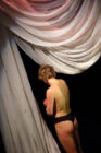 A person stands under dramatic, draped white fabric, wearing black underwear, with their back turned. The lighting creates a contemplative mood.
