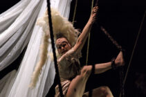 Aerial performer in dramatic pose with white feathers and flowing fabric backdrop. The scene is intense, with focused expression and dynamic movement.