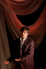 A man in a dark suit stands on stage under dramatic lighting, with red draped curtains in the background. He appears focused and expressive.