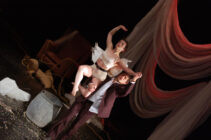 A dancer in a white costume elegantly balances on a man's shoulder on a stage. Draped fabric and scattered props create a dramatic, theatrical ambiance.