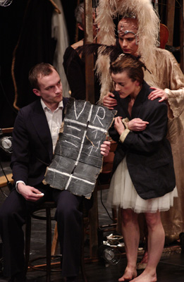 A dramatic stage scene with actors in eclectic costumes. A seated man holds a chalkboard. Emotions are tense, and the mood is intense and theatrical.