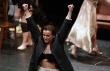 A person in a dark coat raises their arms triumphantly on stage, exuding energy. Blurred background shows people and furniture, suggesting a performance setting.