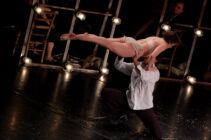 A dancer in a black outfit and white shirt lifts a woman in beige attire horizontally on a dimly lit stage, conveying grace and strength.
