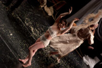 A dancer in beige attire performs expressively on a dimly lit, confetti-strewn stage. A second dancer in flowing robes moves energetically behind them.