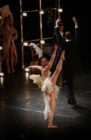 Ballet dancer in a feathered costume performs a high leg lift on stage, surrounded by elegantly dressed individuals, evoking grace and sophistication.