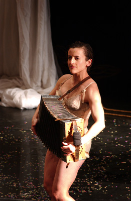 A performer in beige attire plays an accordion on a dimly lit stage. Confetti surrounds them, and a white curtain hangs in the background, creating a theatrical atmosphere.