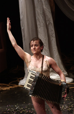 Performer in minimal costume plays an accordion on stage, with a white drape as backdrop. Confetti scattered on the floor, conveying a festive mood.
