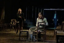 A dimly lit stage shows a tense scene with three people. A woman sits writing on paper at a table with chairs around, while another stands nearby reaching towards her. A third person stands in the shadows, watching. The mood is sombre and dramatic.