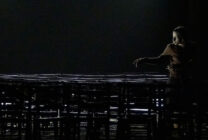 A person stands under dim lighting, reaching out towards empty, stacked chairs in a dark, spacious room, conveying solitude and introspection.