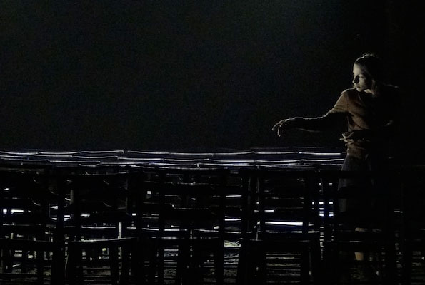 A person stands under dim lighting, reaching out towards empty, stacked chairs in a dark, spacious room, conveying solitude and introspection.