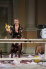 A woman in a floral dress holds a flaming object in a disheveled room. The table is cluttered; her expression is surprised and tense.