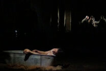 A dimly lit scene shows a person slumped over the edge of a bathtub in the foreground. In the background, four people sit around a table under soft light, creating a sombre mood.