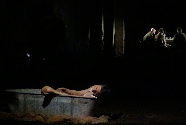A dimly lit scene shows a person slumped over the edge of a bathtub in the foreground. In the background, four people sit around a table under soft light, creating a sombre mood.