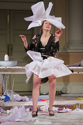 A woman in a black robe joyfully tosses white paper into the air in a cluttered room, expressing chaos and creativity. Papers and art supplies cover the floor.