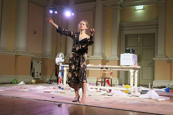 A woman in a floral robe stands on a cluttered stage with scattered papers and objects, arms raised in an expressive, dramatic pose. The setting is theatrical.