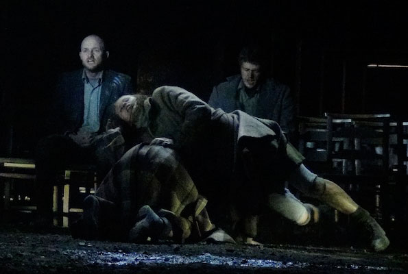 A dimly lit scene shows a sombre mood. Three men sit on scattered chairs; one leans over another wrapped in a checked blanket, suggesting comfort or despair.