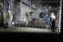 Dark, industrial setting with people in coats jumping over rows of chairs amid bright stage lights, creating a sense of urgency and chaos.