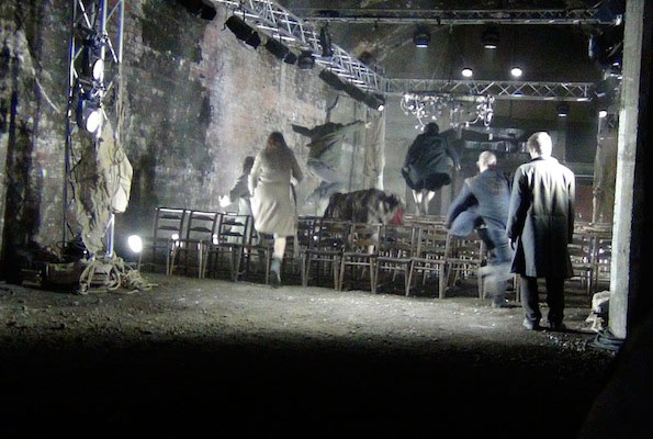 Dark, industrial setting with people in coats jumping over rows of chairs amid bright stage lights, creating a sense of urgency and chaos.