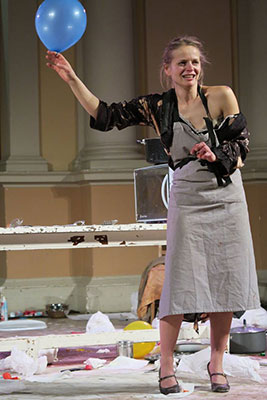 A woman in a tattered outfit holds a blue balloon, standing amidst a messy room with scattered papers and objects, conveying a lively, chaotic scene.