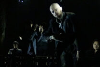 A group of five people dressed in dark clothing stand in a dimly lit space. The atmosphere is tense, with a spotlight on a bald man in the foreground.