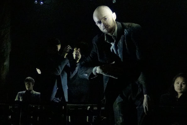 A group of five people dressed in dark clothing stand in a dimly lit space. The atmosphere is tense, with a spotlight on a bald man in the foreground.