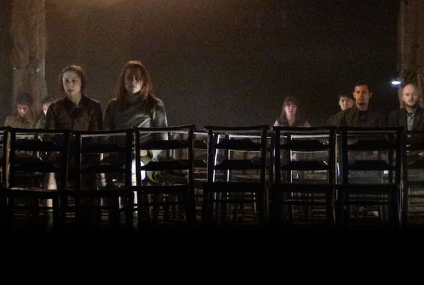 A group of people stands solemnly behind a row of empty chairs in a dimly lit setting. The atmosphere is tense and serious, conveying anticipation.