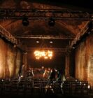 Dimly lit underground venue with exposed brick walls, metal beams, and a central chandelier. A few people are arranging chairs, creating an intimate, moody atmosphere.