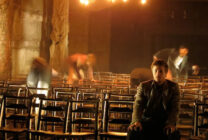 A dimly lit room with rows of wooden chairs, one person sitting calmly in the foreground while blurred figures in motion suggest hurried activity. The atmosphere feels tense and dramatic.