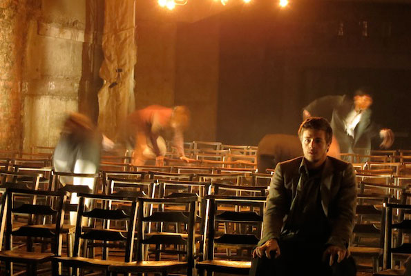 A dimly lit room with rows of wooden chairs, one person sitting calmly in the foreground while blurred figures in motion suggest hurried activity. The atmosphere feels tense and dramatic.