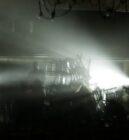 A musician plays drums under dramatic stage lighting, silhouetted by bright beams and shadowy surroundings, creating a mysterious and intense atmosphere.