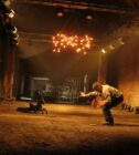 Dimly lit warehouse with dirt floor and scattered chairs in background. A person lunges forward toward a small, dark horse figurine under glowing chandelier.