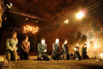 Seven people kneel in a dimly lit, rustic room with exposed brick walls, under dramatic lighting. The mood is solemn and introspective.