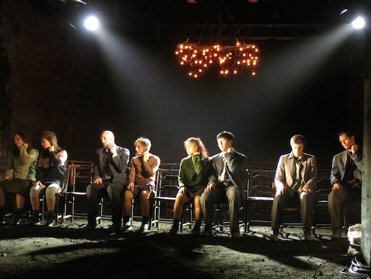 A group of people sit in a row on chairs under dim stage lights, with an illuminated sign above. The scene conveys a dramatic and introspective mood.