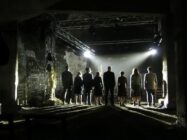 A group of people, silhouetted and standing in a line, face forward in a dimly lit industrial setting. Overhead lights cast dramatic shadows.