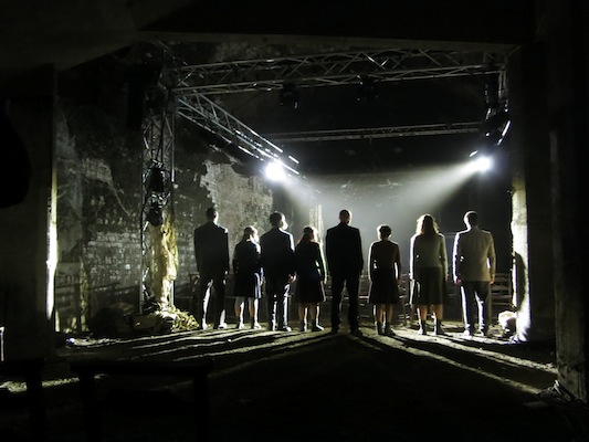 A group of people, silhouetted and standing in a line, face forward in a dimly lit industrial setting. Overhead lights cast dramatic shadows.