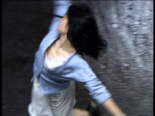 Woman with flowing dark hair, dressed in a light blue sweater and white dress, spins joyfully on a wet pavement, conveying a sense of freedom.