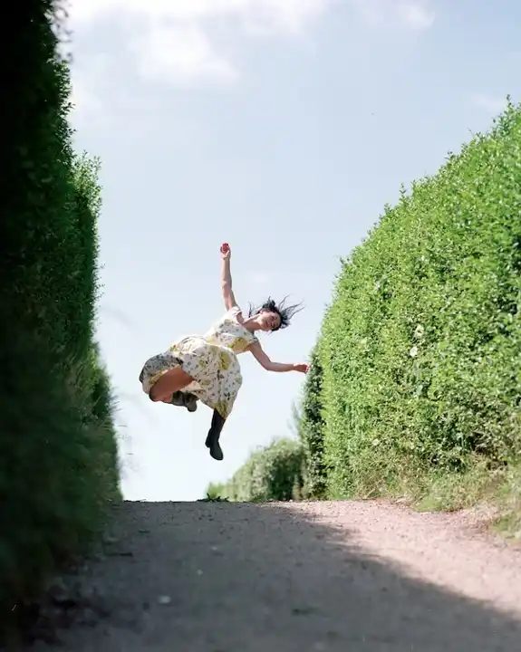 A woman in a floral dress joyfully leaps between tall green hedges on a sunny day, arms and legs outstretched, capturing a moment of exuberance.