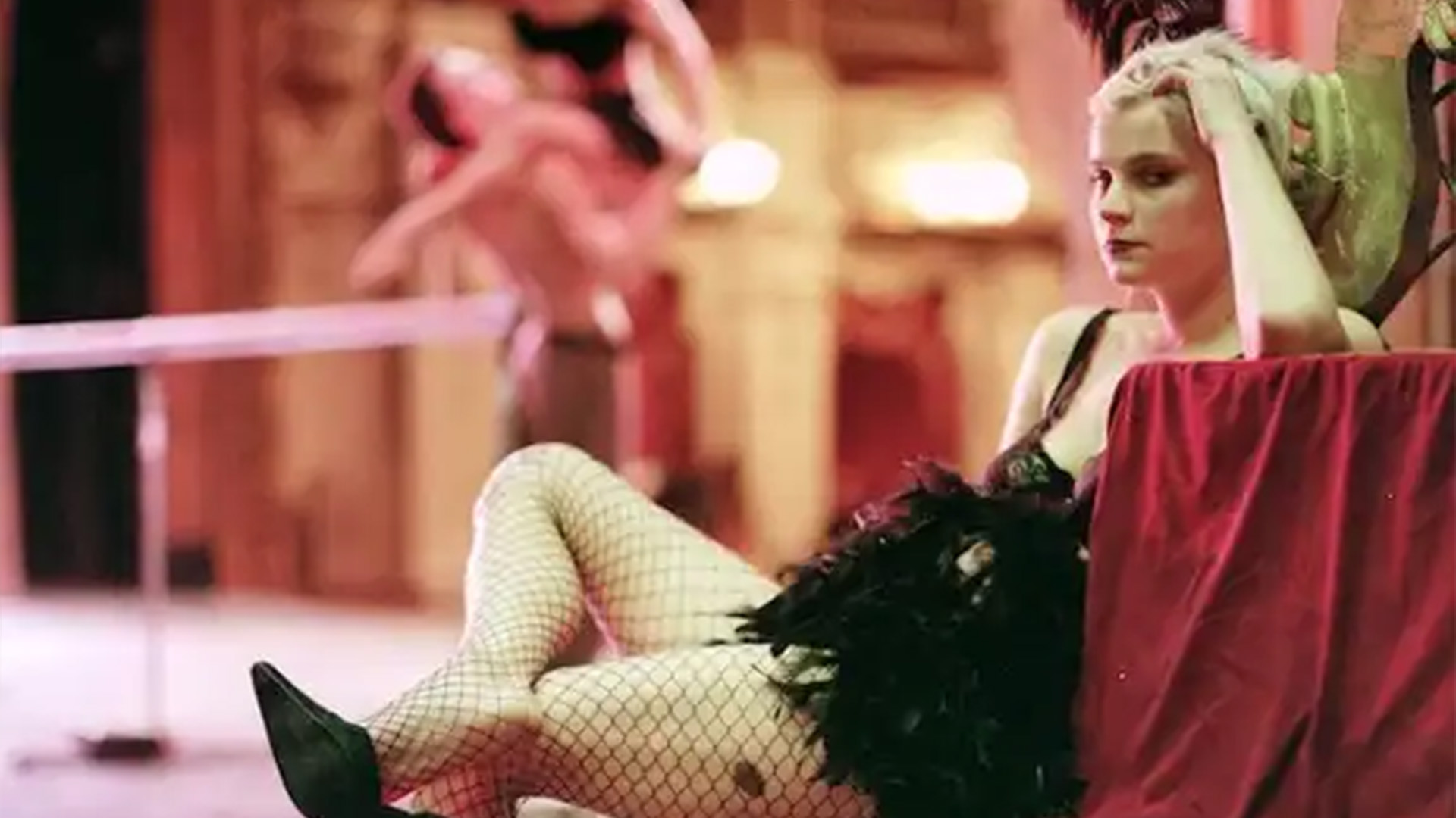 Young woman in fishnet stockings and black dress sits pensively backstage, with dancers performing energetically in the softly lit background.