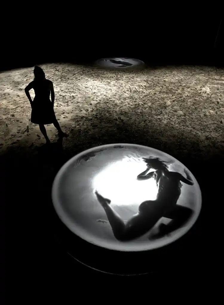 Silhouette of a woman standing on a textured surface, looking at a large lit circular platform. A floating, shadowy figure is visible inside it, creating a mysterious, contemplative mood.