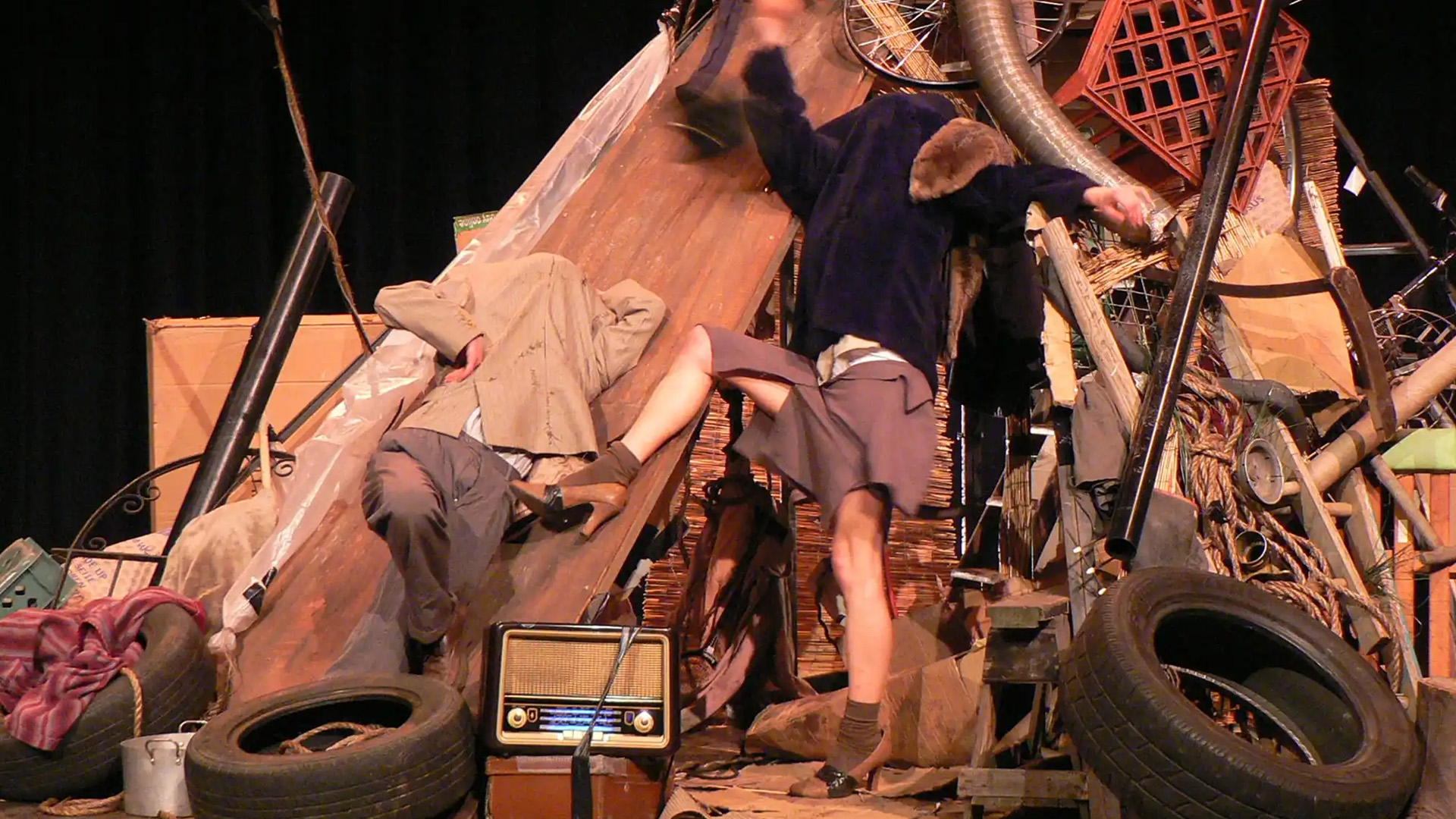 Two people amidst a cluttered pile of junk, including tires and an old radio, create a chaotic scene. Their dynamic poses suggest movement and instability.