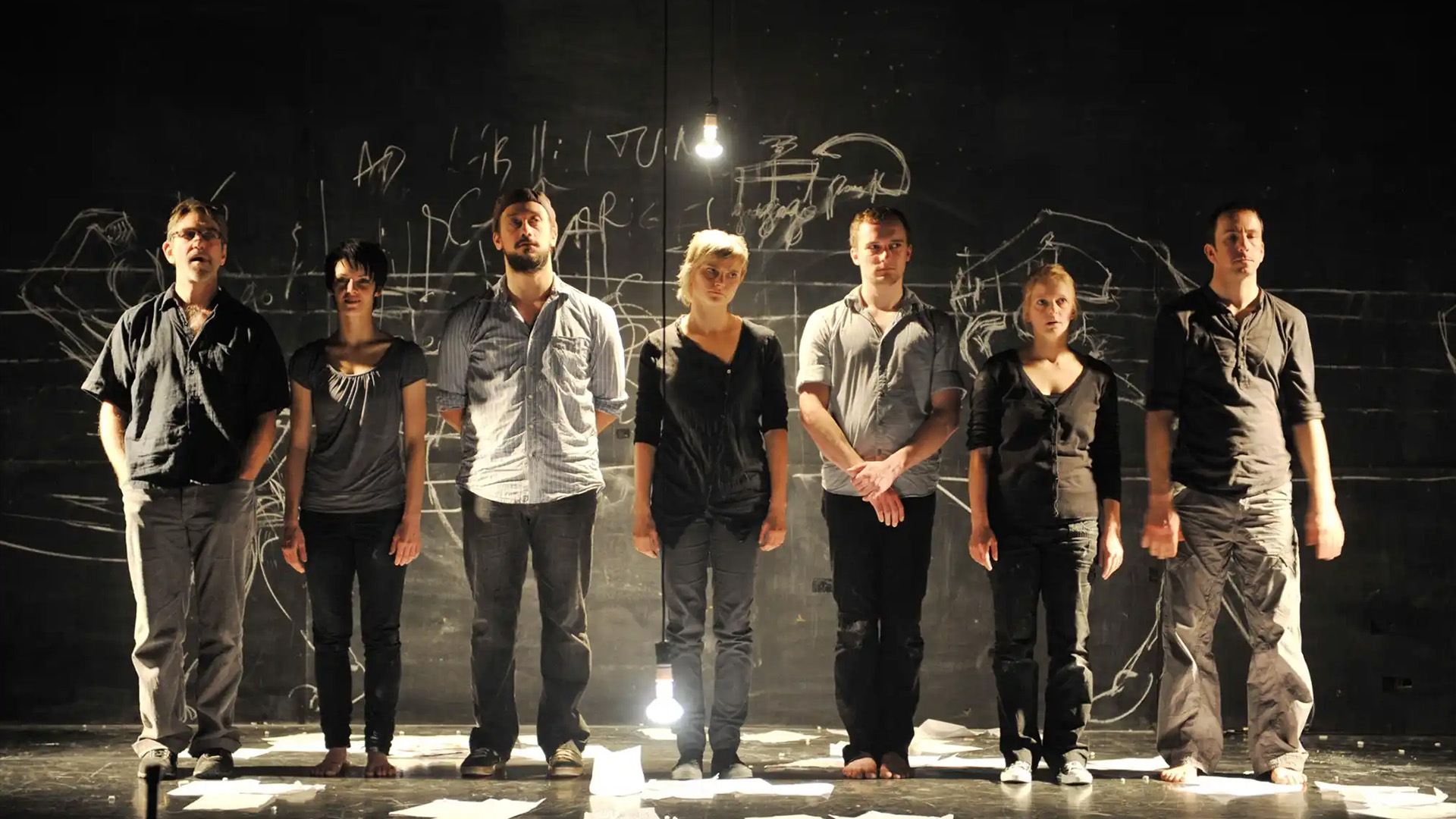 Seven people stand in a row under dim lighting on a stage with chalk drawings on the blackboard behind them. The atmosphere is serious and introspective.