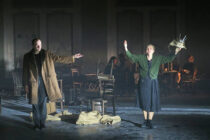 A theatrical scene with two actors on a dimly lit stage. One wears a long coat, the other raises her arms, holding a basket, conveying drama and tension.